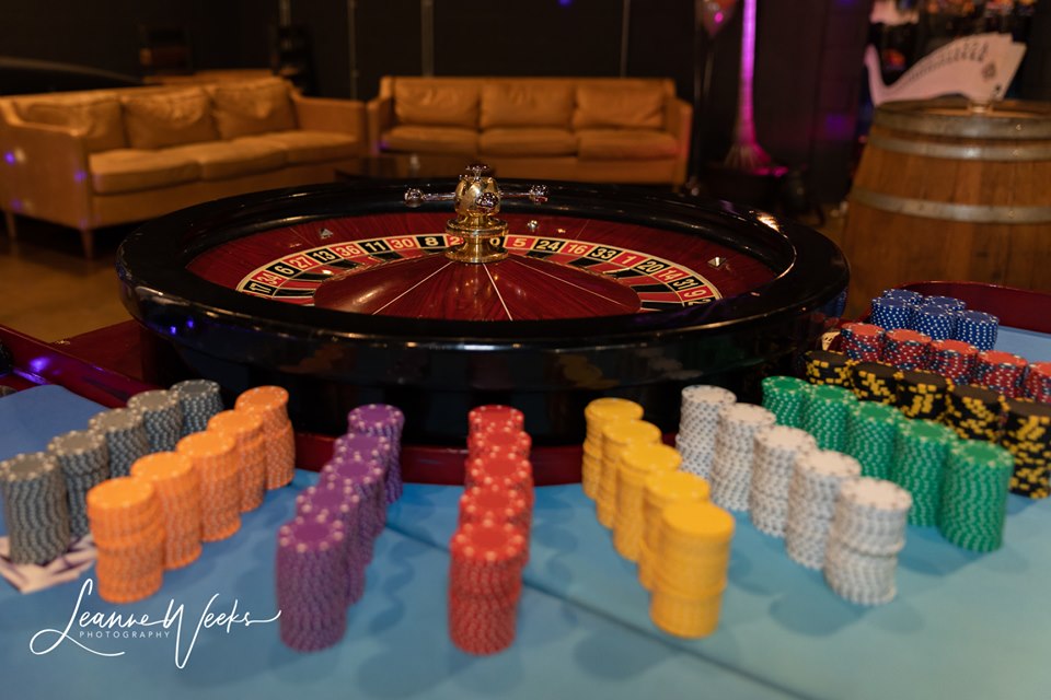 A close-up of a casino roulette table with a spinning wheel and neatly stacked poker chips in various colours. In the background, there are brown leather sofas and a wooden barrel.